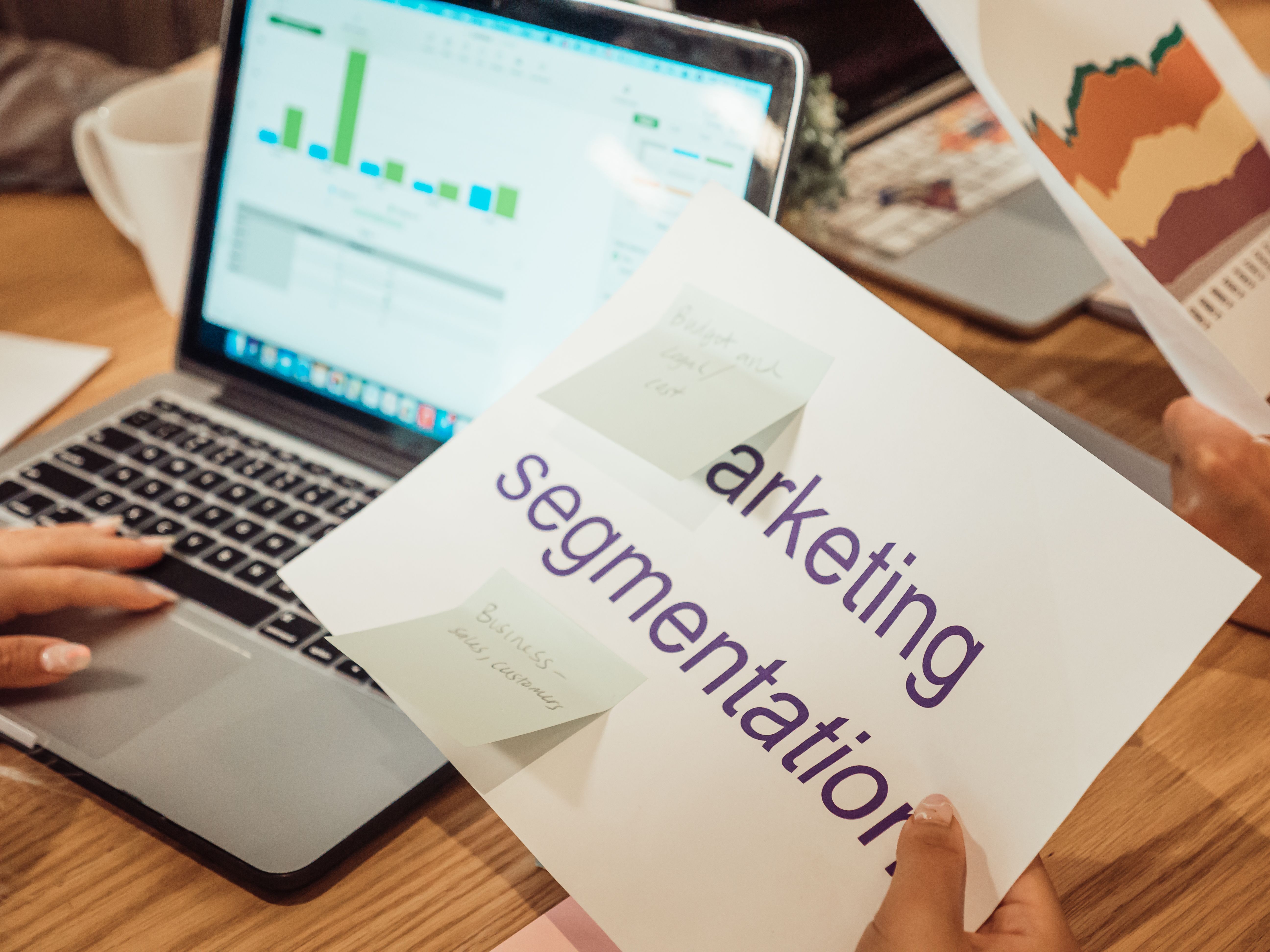 Photo illustration of hands reviewing marketing charts on a laptop and printed documents in a modern workspace