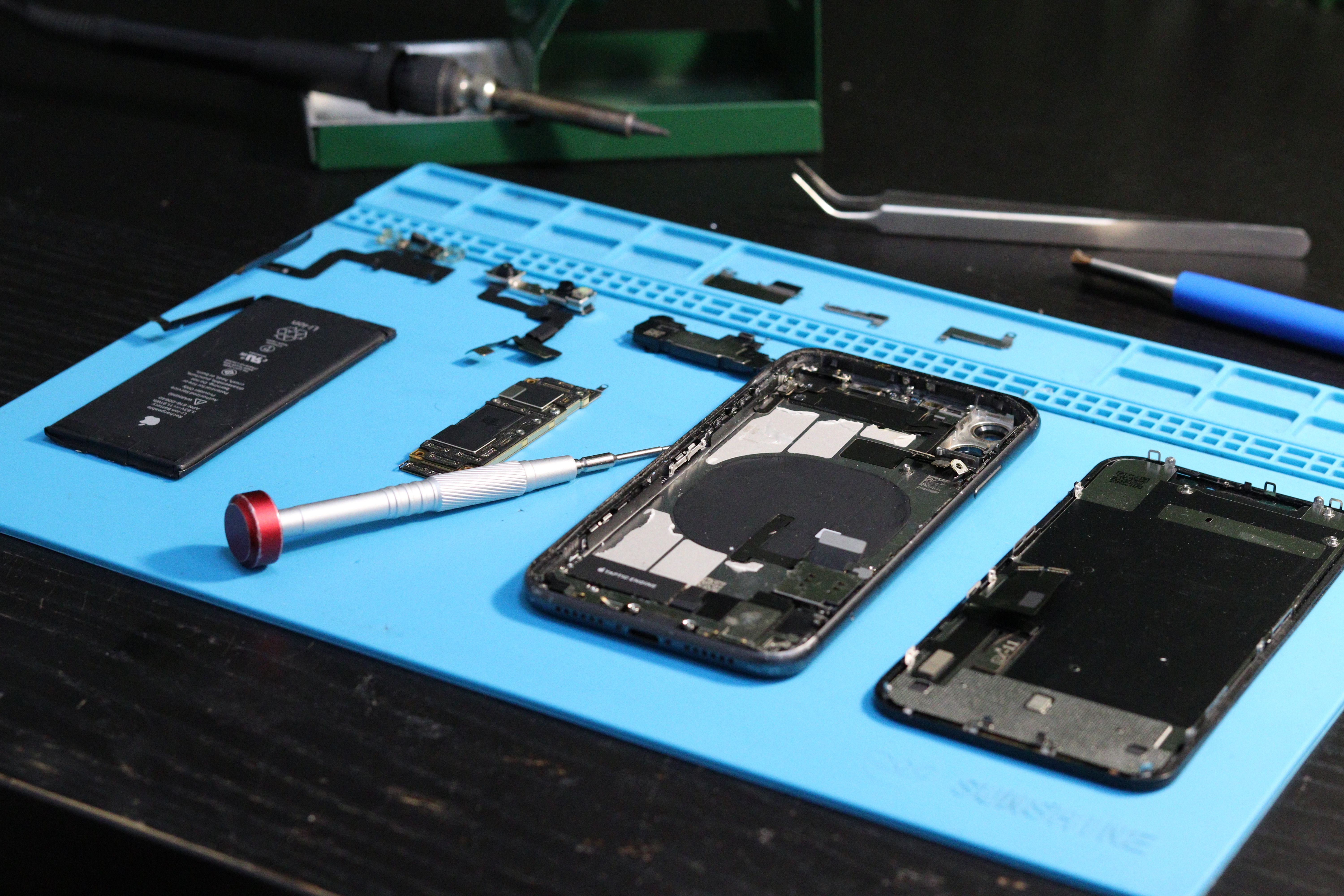 Close-up of disassembled smartphone components, circuit boards, and precision tools on a blue technician workbench.