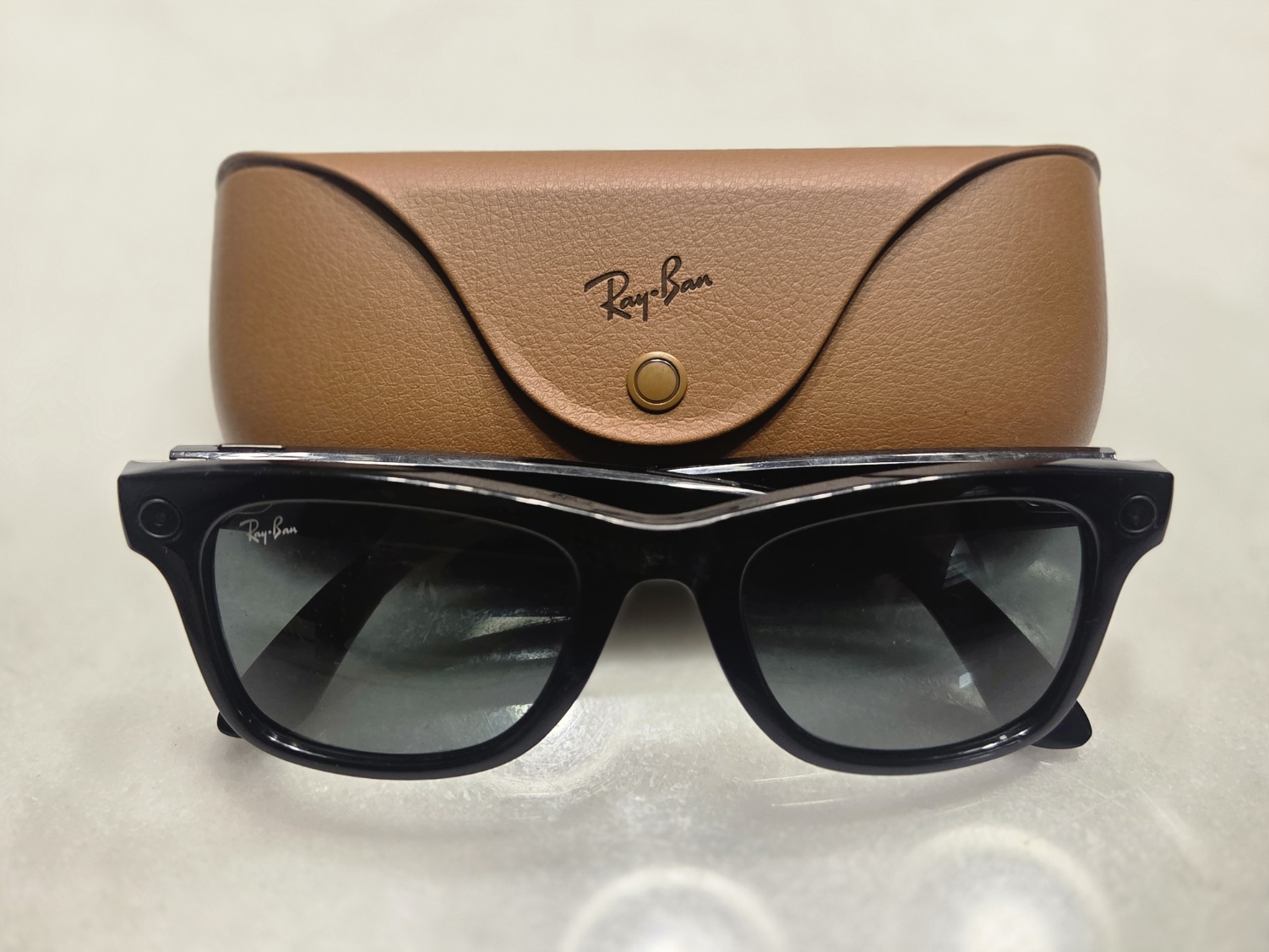 Photograph of black Ray-Ban Meta Gen 1 smart glasses positioned in front of their brown leather charging case.