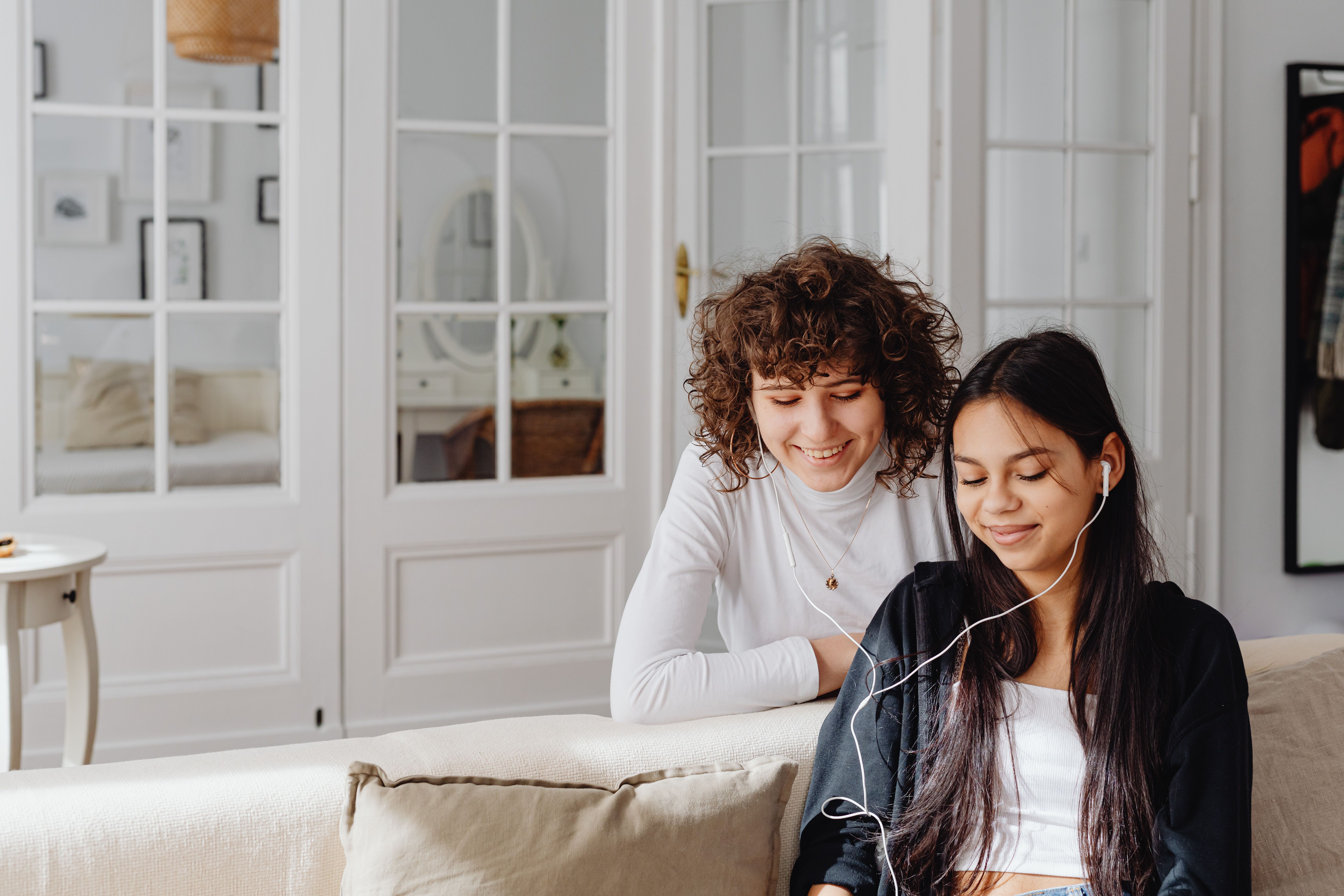 Two friends relaxing on a living room sofa, sharing headphones and listening to music together.